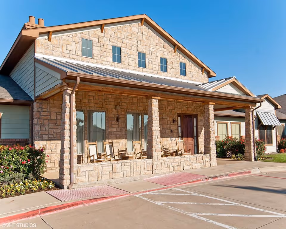 Exterior view of Walnut Creek Assisted Living and Memory Care building with stone facade, a covered porch with wooden rocking chairs, and landscaped flower beds under a clear blue sky.