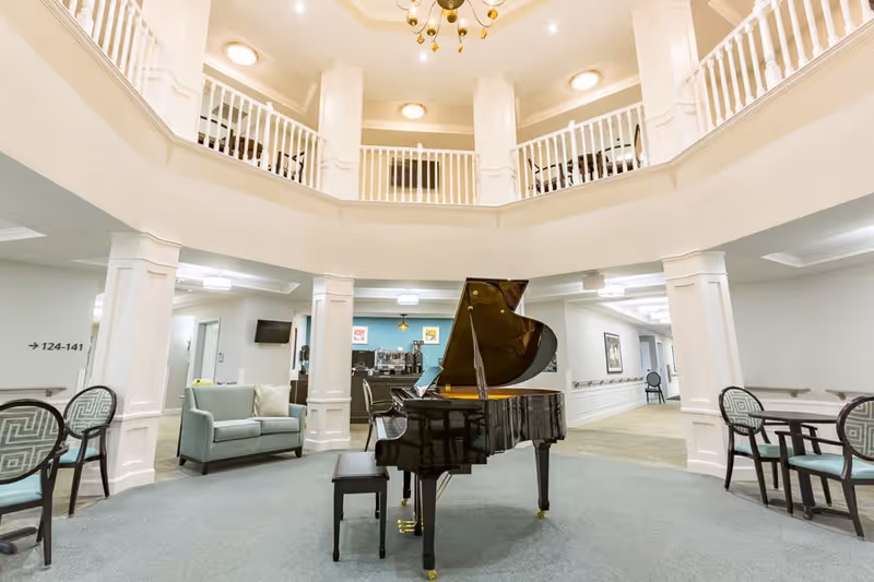 Interior view of a senior living facility common area featuring a black grand piano in the center, surrounded by seating including chairs and a small sofa. The space has white pillars, a two-story ceiling with a balcony railing above, and soft lighting. There is a hallway visible in the background with directional signage.