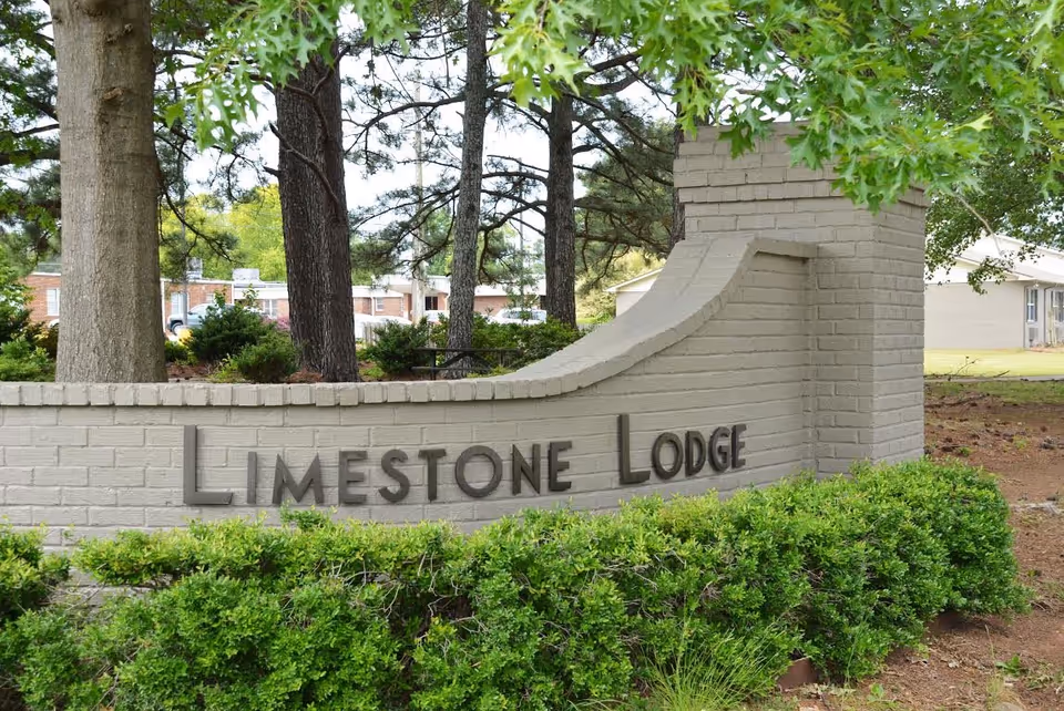 A beige brick sign with the words 'Limestone Lodge' mounted on it, surrounded by green bushes and trees, with buildings visible in the background.