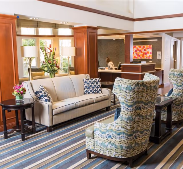 A bright and welcoming living room area in a senior living facility featuring a beige sofa with patterned cushions, two patterned armchairs, a striped carpet, side tables with flower arrangements, and a reception desk in the background where a person is seated working on a computer.