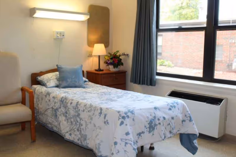 A single bed with floral patterned bedding and a blue pillow in a small, tidy room. Next to the bed is a wooden nightstand with a lamp and a flower arrangement. A beige armchair is positioned to the left of the bed. A large window with blue curtains lets in natural light, showing a brick building outside.