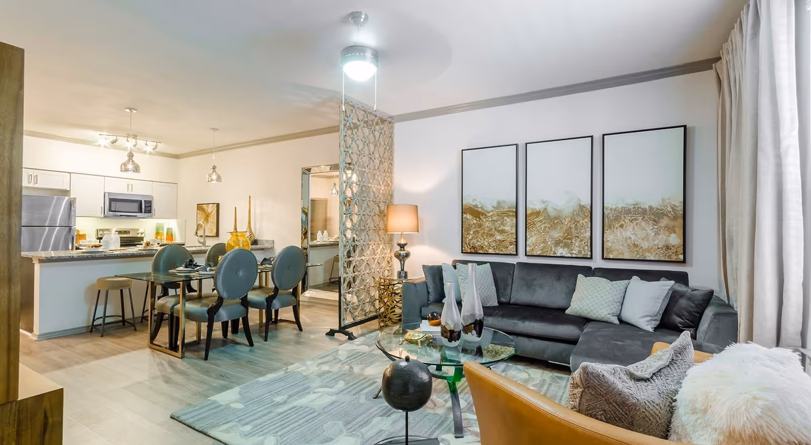 A modern open-concept living and dining area with a gray sectional sofa, glass coffee table, and three abstract paintings on the wall. The dining area has a table set with four chairs and is adjacent to a kitchen with stainless steel appliances and white cabinetry. The space is well-lit with ceiling lights and a floor lamp, and features light wood flooring and neutral-colored walls.