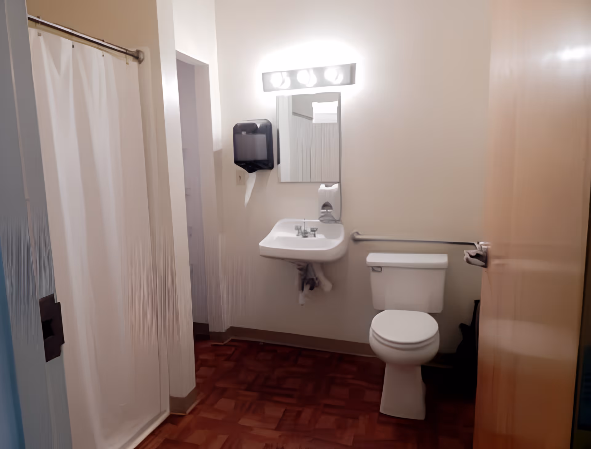 Small accessible bathroom with a shower curtain, wall-mounted sink and mirror, toilet with a grab bar, and a wall-mounted paper towel dispenser.