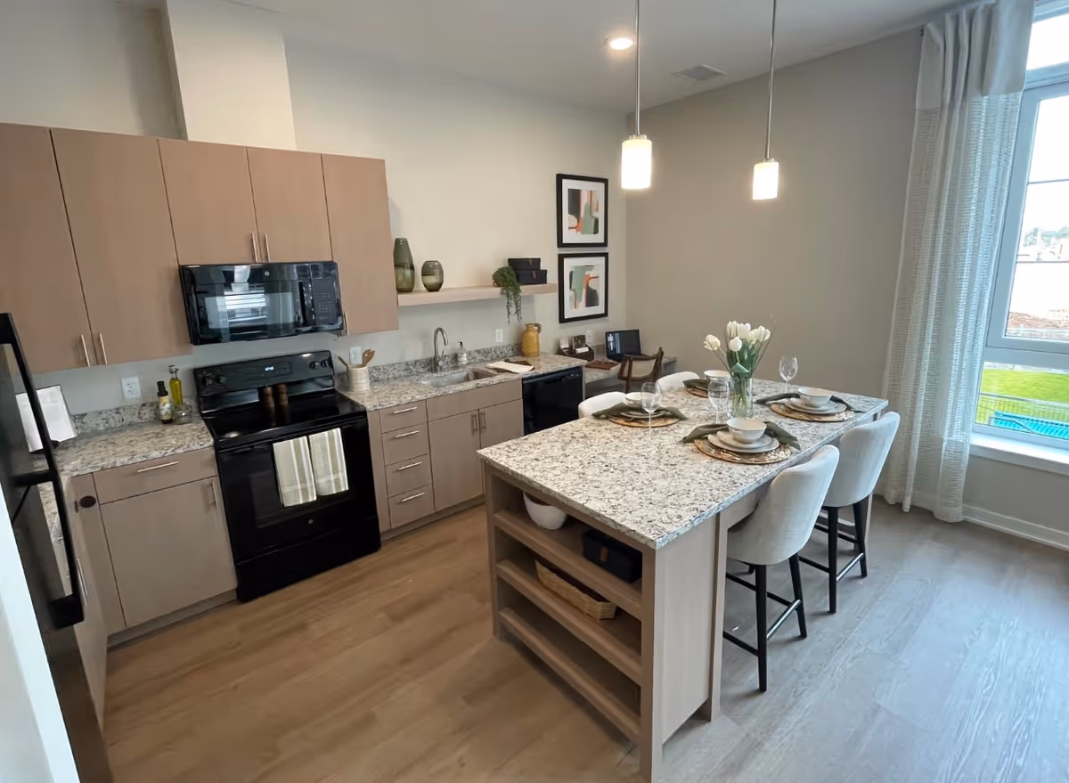 Modern kitchen with light wood cabinets, black appliances including a stove and microwave, granite countertops, and a kitchen island set with four place settings and white upholstered chairs. The room has light wood flooring, two pendant lights hanging above the island, and a large window with sheer curtains letting in natural light.