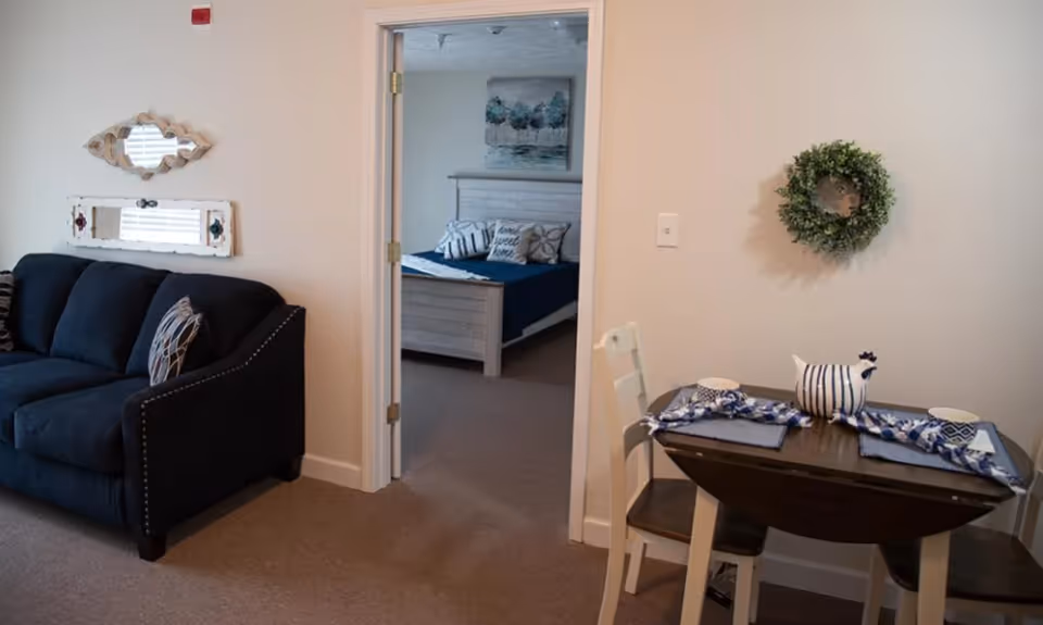 Interior view of a senior living facility showing a small dining area with a wooden table set for two, a dark blue couch on the left, and an open doorway leading to a bedroom with a bed, pillows, and wall art.