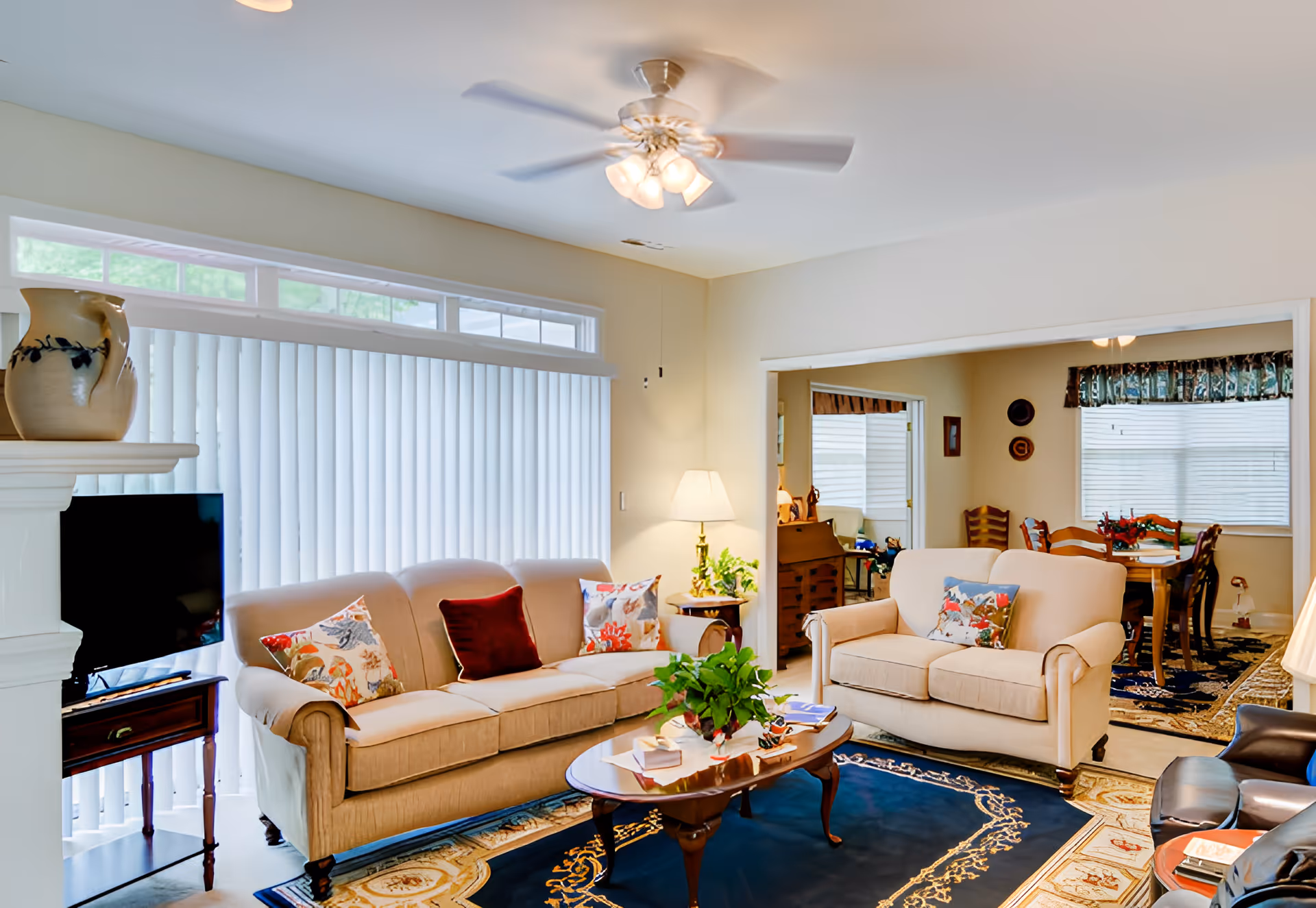 A cozy living room with beige sofas adorned with colorful cushions, a wooden coffee table with a plant and books, a ceiling fan with lights, a TV on a small wooden stand, and a lamp on a side table. The room opens into a dining area with a wooden dining table and chairs, decorated with a floral centerpiece and window blinds.