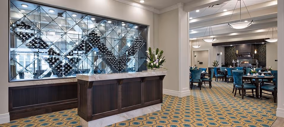 Interior view of a senior living facility dining area featuring a decorative wine rack with bottles displayed in a geometric pattern behind a wooden counter. The room has patterned carpet flooring, teal upholstered chairs around dining tables, and modern ceiling lights. There are floral arrangements on the counter and tables, and a fireplace is visible in the background.