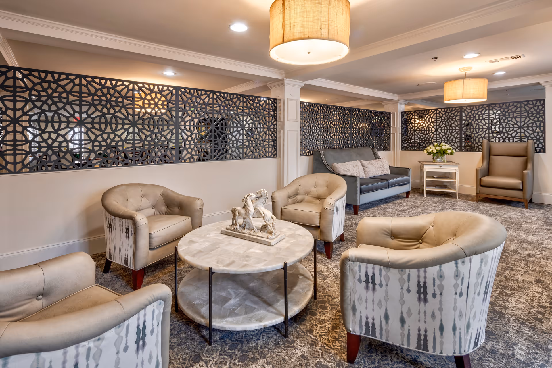 A cozy seating area in a senior living facility with four beige patterned armchairs arranged around a round marble coffee table with a decorative sculpture. In the background, there is a gray sofa with two cushions, a side table with a flower arrangement, and a high-back armchair. The room features decorative black lattice panels on the walls and warm overhead lighting with large drum pendant lamps.