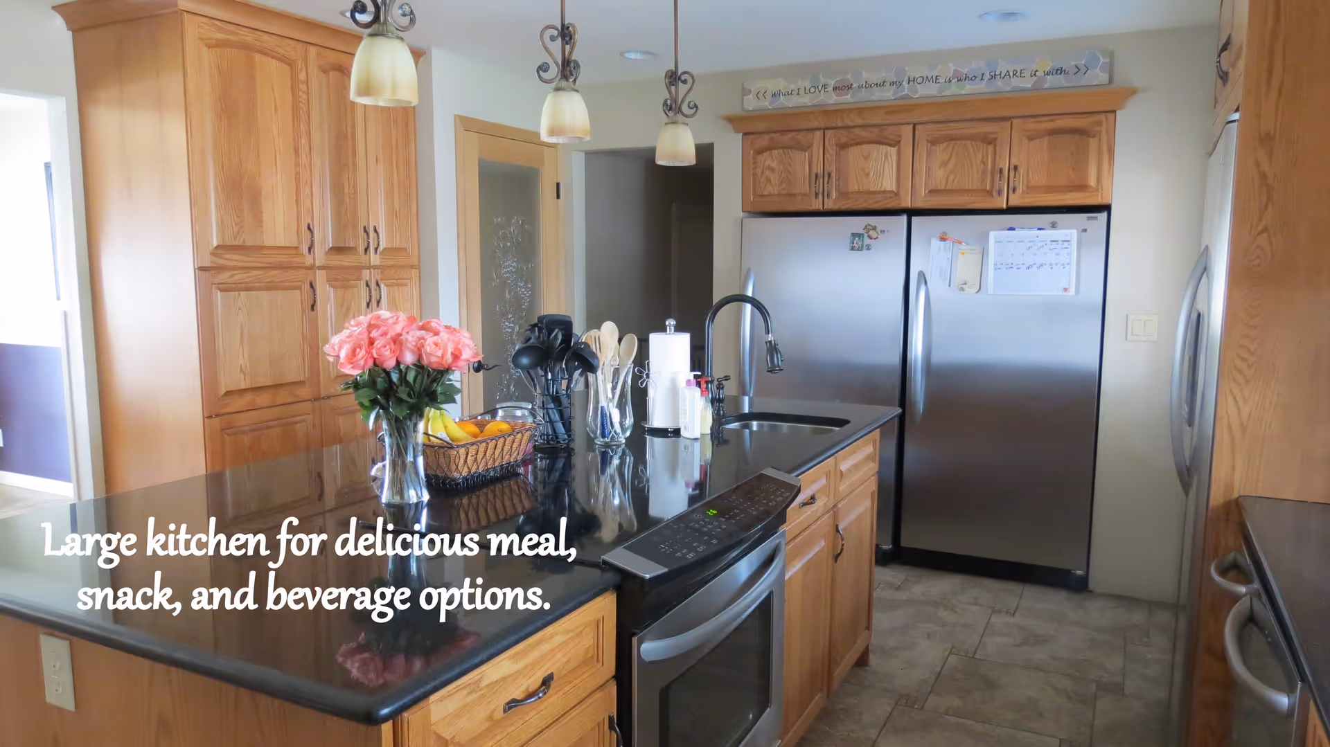 A large kitchen with wooden cabinets, a black countertop island with a vase of pink roses, a basket of fruit, kitchen utensils, and a paper towel holder. Stainless steel refrigerator and oven are visible, along with three pendant lights hanging from the ceiling. A decorative sign above the refrigerator reads, 'What I LOVE most about my HOME is who I SHARE it with.'