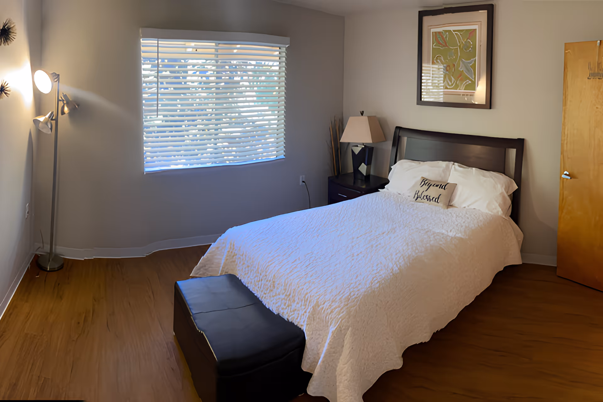 A bedroom with a bed covered in a white quilt and pillows, one pillow has the words 'Beyond Blessed'. There is a black cushioned bench at the foot of the bed, a nightstand with a lamp, a framed artwork on the wall above the bed, a window with blinds letting in daylight, a wooden door, and a floor lamp with three lights on the left side.