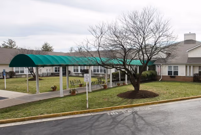 Exterior view of a senior living facility with a covered walkway supported by white columns and a green canopy. There are benches under the canopy, a tree with no leaves in a mulched area, and a building with multiple windows and a chimney in the background. The pavement in front has a 'NO PARKING' marking.