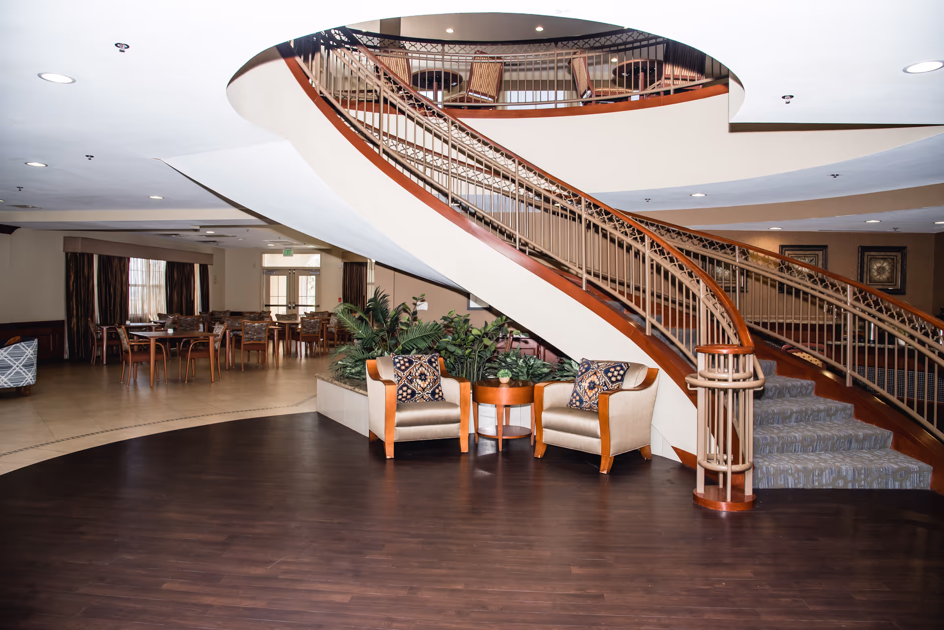 Interior view of a senior living facility featuring a curved staircase with ornate railings. In the foreground, there are two cushioned armchairs with patterned pillows and a small round wooden table between them. Behind the seating area, there is a planter with green plants. In the background, there is a dining area with multiple wooden tables and chairs, and large windows with curtains allowing natural light to enter.