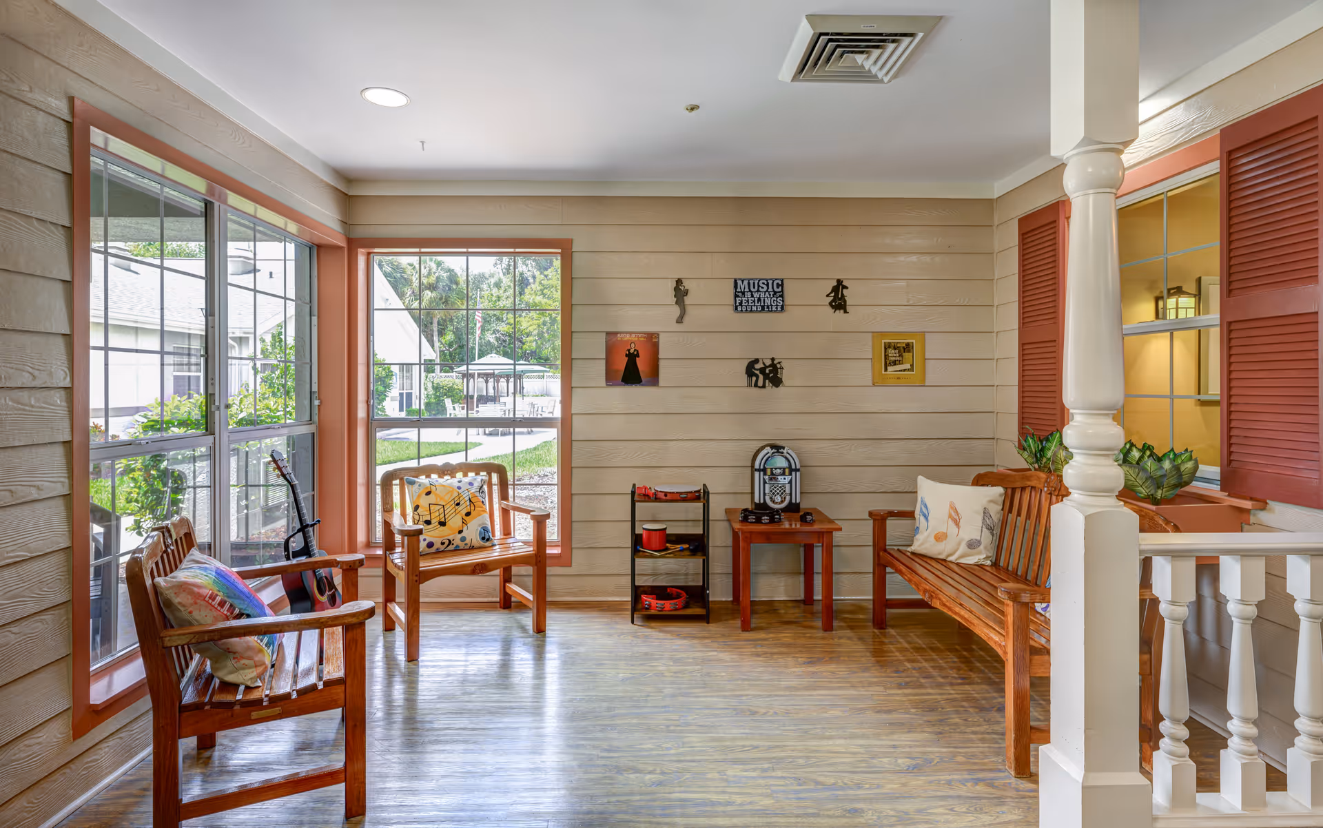 A bright and cozy sitting area with wooden benches and chairs adorned with colorful cushions featuring musical notes. The room has large windows with a view of a garden and outdoor gazebo. The walls are decorated with music-themed artwork and a small jukebox sits on a wooden table. The floor is wooden, and the space has a warm, inviting atmosphere.