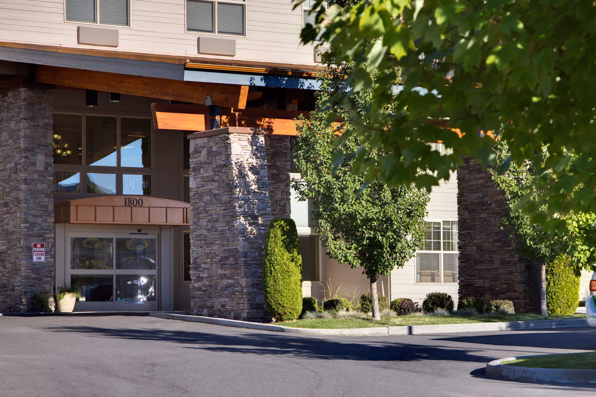 Entrance of a senior living facility with stone pillars, large windows, and a covered drop-off area. The building number 1800 is displayed above the automatic glass doors. Trees and shrubs are planted around the entrance, and a 'No Parking Loading Zone' sign is visible on the left side.