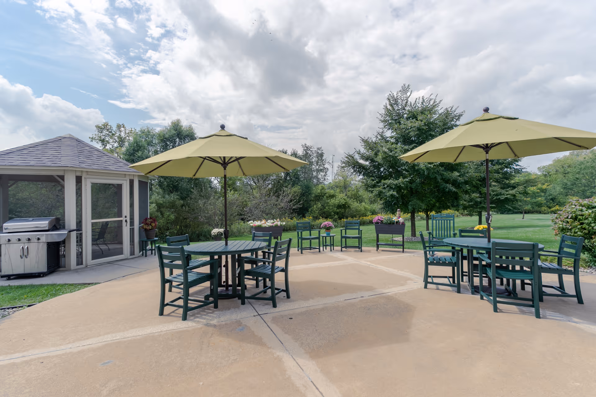 Outdoor patio area with two round tables each surrounded by green chairs and shaded by large green umbrellas. There is a small screened gazebo with a grill on the left side and various potted plants and flowers around the patio. The background features green trees and a partly cloudy sky.