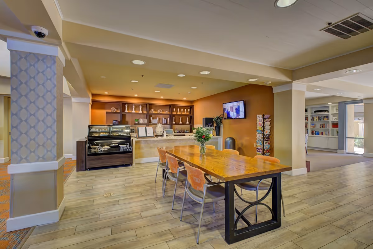 Interior view of a senior living facility common area with a wooden table and six chairs, a display case with baked goods, a counter with decorative items, a wall-mounted TV, and a rack of brochures. The space has tiled flooring, warm lighting, and an open layout leading to a bookshelf area.