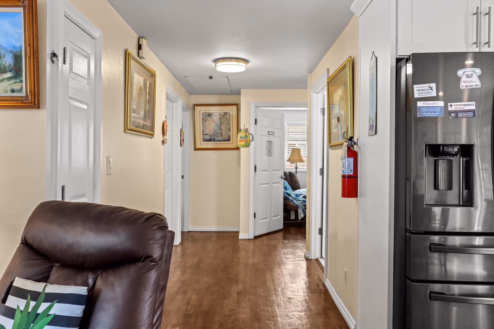 View down a hallway in a senior living facility with light yellow walls and wooden flooring. On the left, there is a brown leather chair with a striped pillow. Several framed paintings hang on the walls. At the end of the hallway, an open door reveals a room with a lamp and a bed covered with a blue blanket. On the right side, part of a stainless steel refrigerator with magnets and a fire extinguisher mounted on the wall are visible.