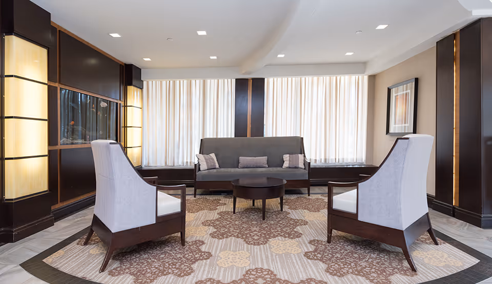 A modern lounge area with two high-back armchairs and a sofa arranged around a round coffee table on a patterned carpet. The room features large windows with sheer curtains, warm lighting fixtures on the walls, and a framed artwork on one side.