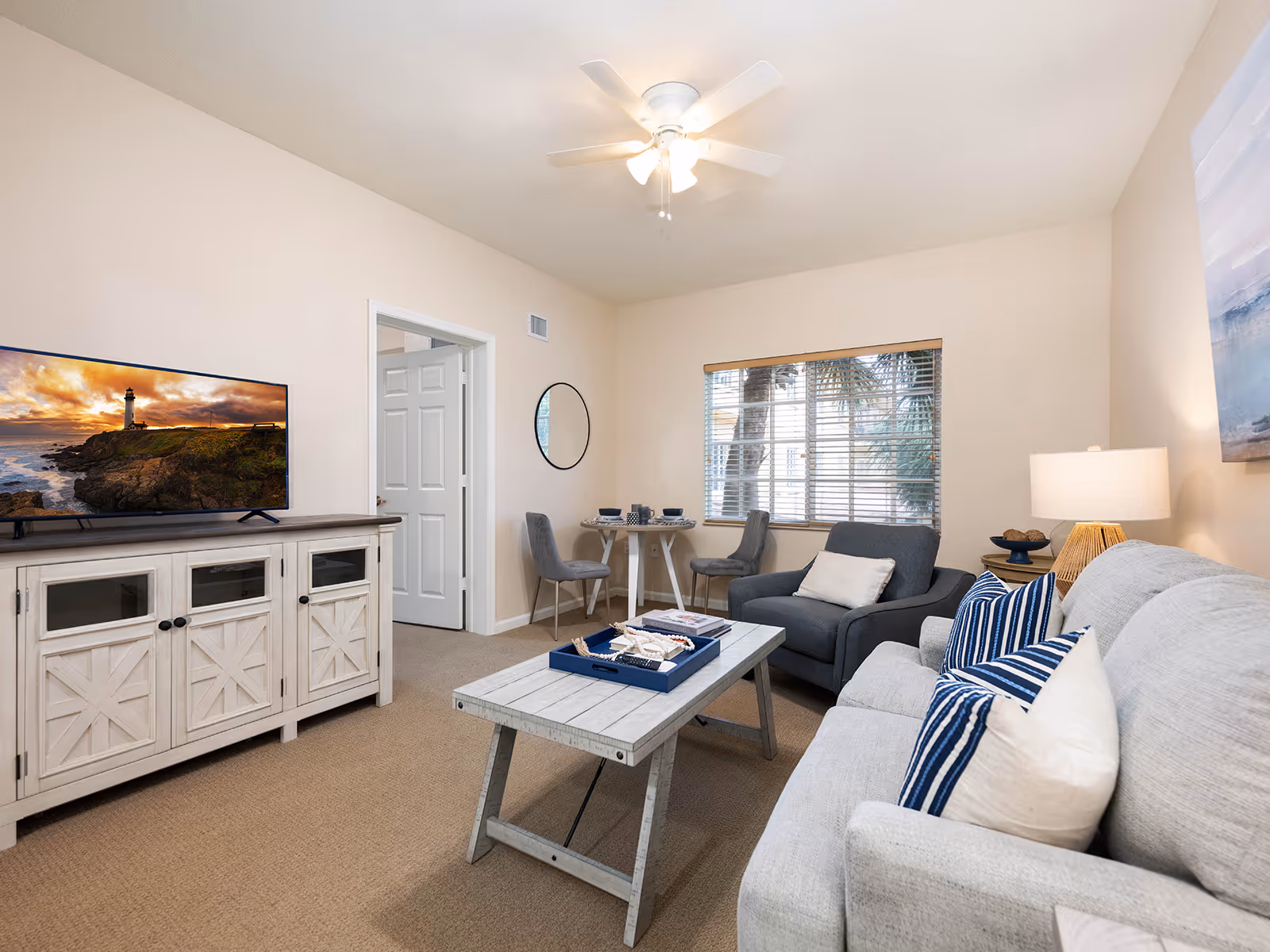 A cozy living room with a light gray sofa adorned with blue and white striped pillows, a matching armchair, and a rustic wooden coffee table. A white cabinet with a flat-screen TV displaying a lighthouse scene is against one wall. A small round dining table with two gray chairs is positioned near a window with blinds. The room is softly lit by a ceiling fan light and a table lamp, with a large painting hanging on the wall.