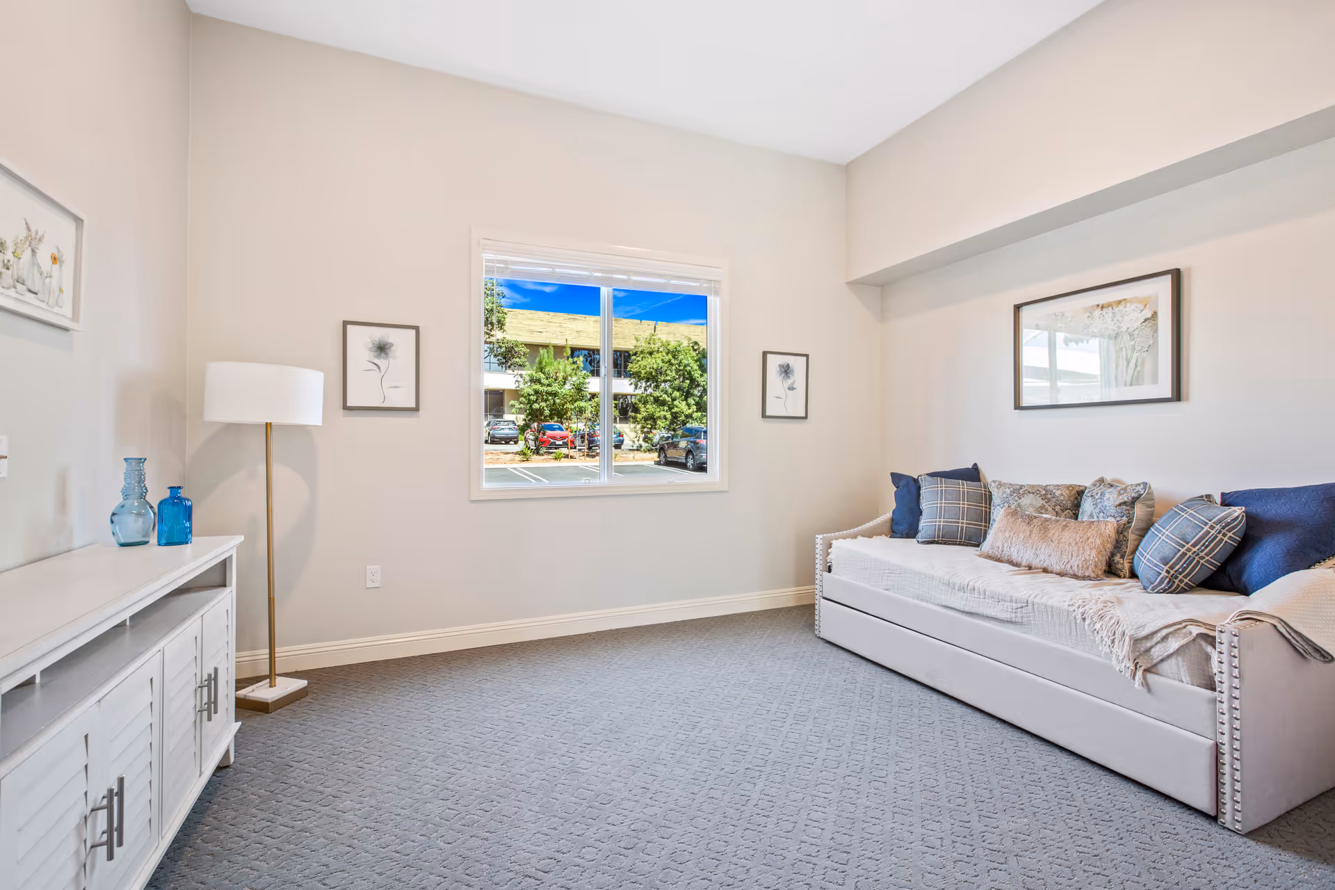 A bright, clean room with a large window showing a view of parked cars and trees outside. Inside, there is a white daybed with multiple decorative pillows and a throw blanket on the right side. On the left side, there is a white cabinet with two blue glass vases on top and a tall floor lamp with a white shade. The walls are light-colored and decorated with framed floral artwork.