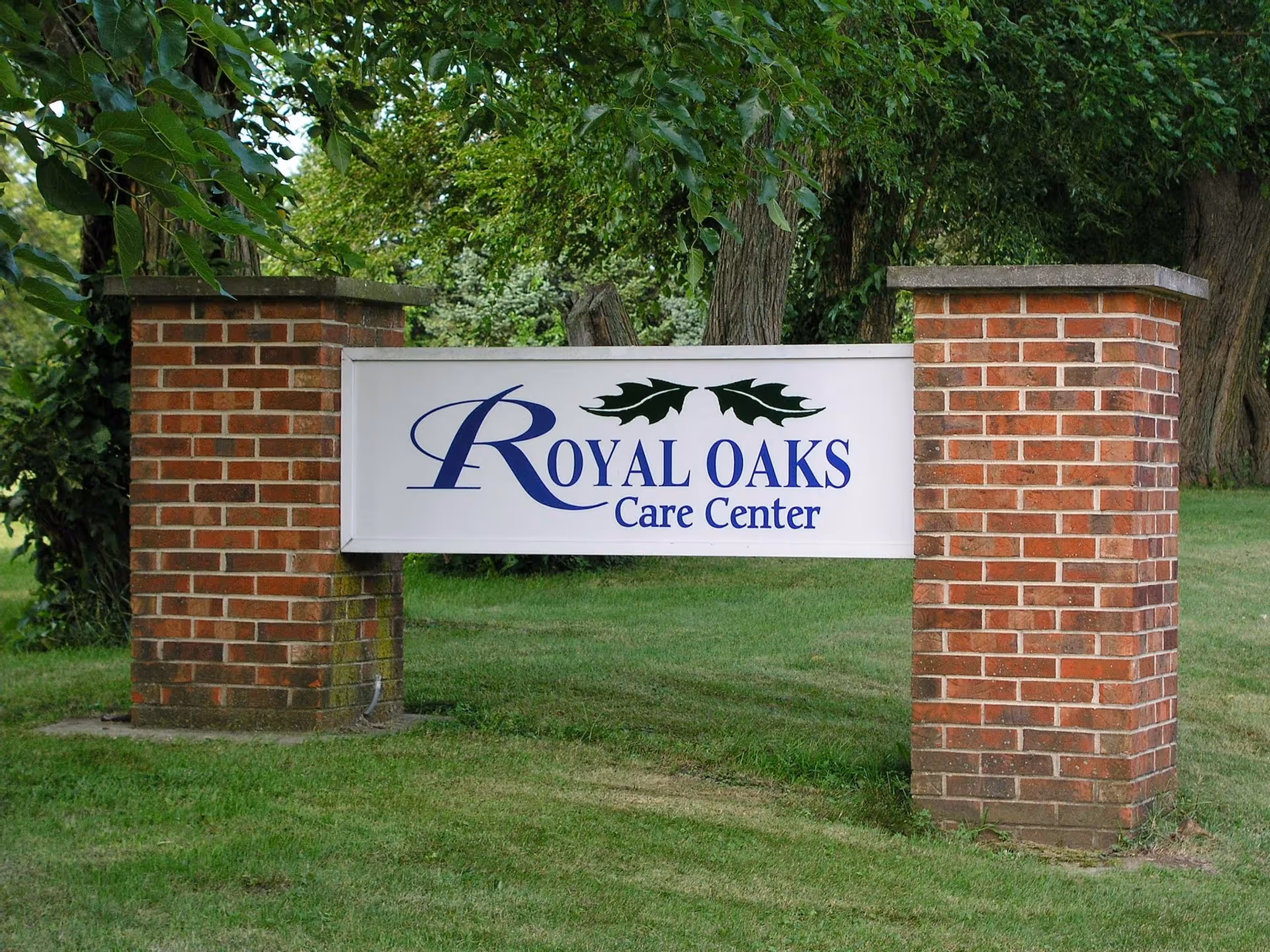 A white sign with blue text reading 'Royal Oaks Care Center' mounted between two brick pillars on a grassy area with trees in the background.