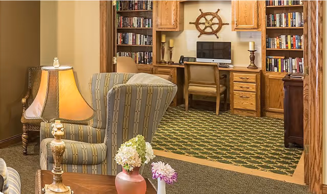 Cozy interior room with a striped upholstered armchair and a wooden side table holding a lamp and flower vases in the foreground. In the background, there is a built-in wooden desk with a computer, a chair, bookshelves filled with books, and a decorative ship wheel mounted on the wall.