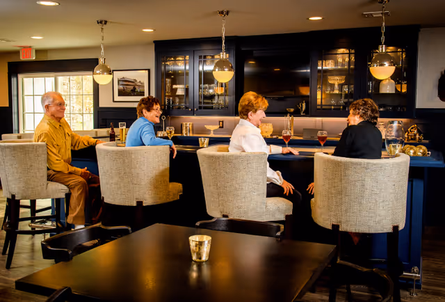 Four senior residents sit on upholstered bar stools chatting at a lounge-style bar with drinks and glass-front cabinets behind them.