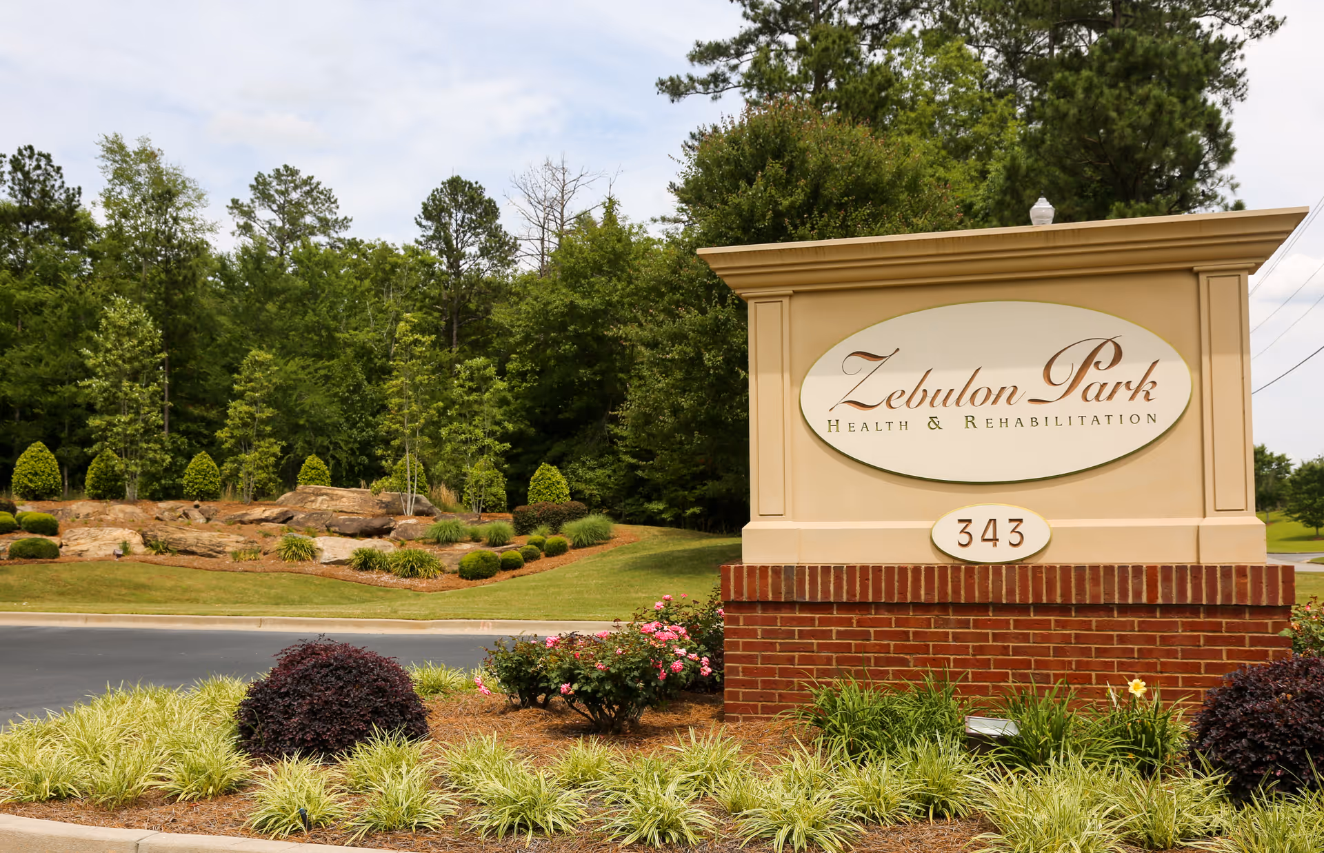 Entrance sign for Zebulon Park Health & Rehabilitation with the address number 343, surrounded by landscaped greenery and flowers, with trees and a partly cloudy sky in the background.
