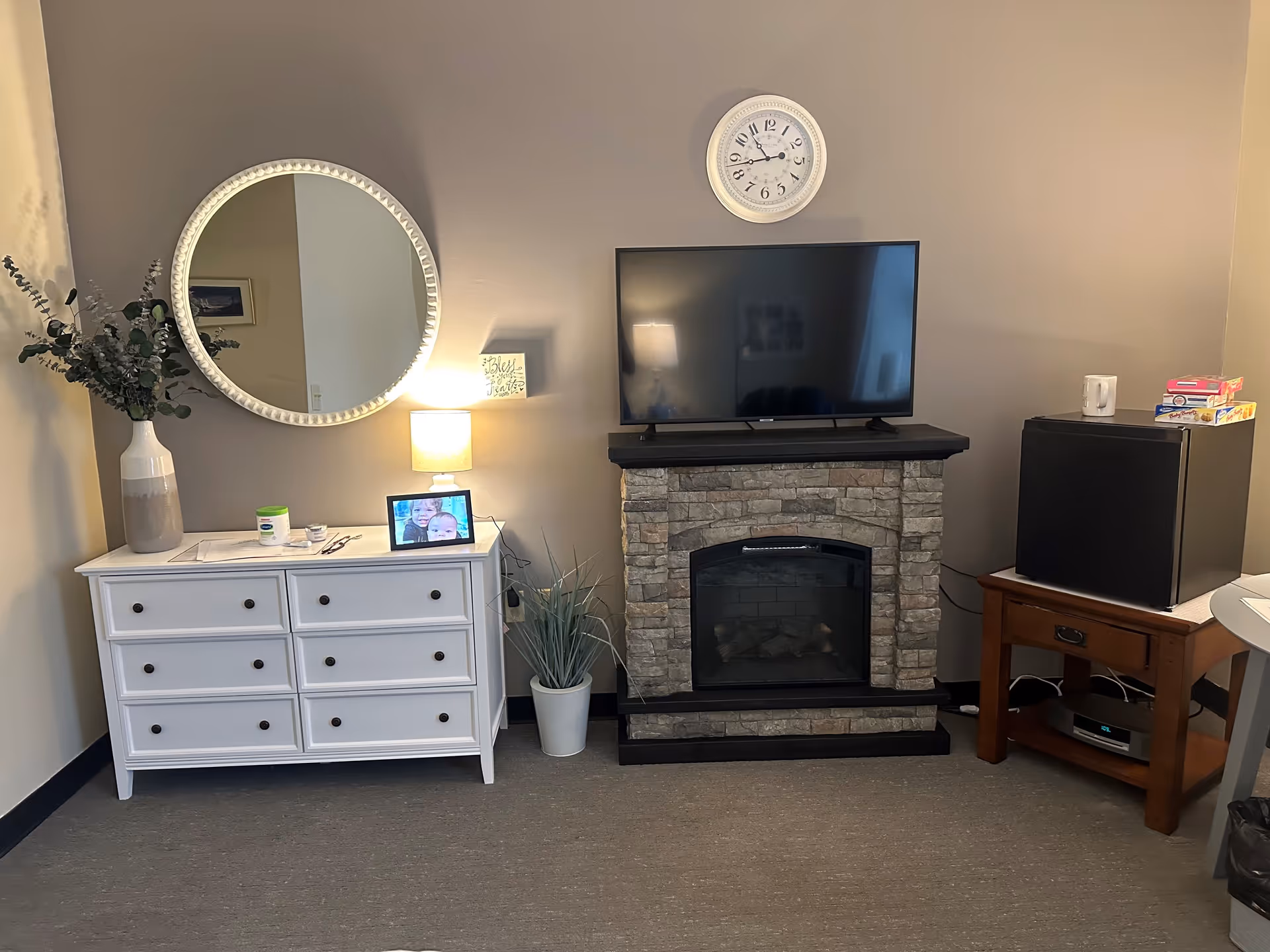 A cozy living area featuring a white dresser with six drawers, a round decorative mirror above it, a small table lamp, a digital photo frame, and a vase with greenery. Next to the dresser is a stone fireplace with a flat-screen TV mounted on top and a white clock on the wall above the TV. To the right, there is a small wooden table holding a black mini refrigerator with a mug and some board games on top.