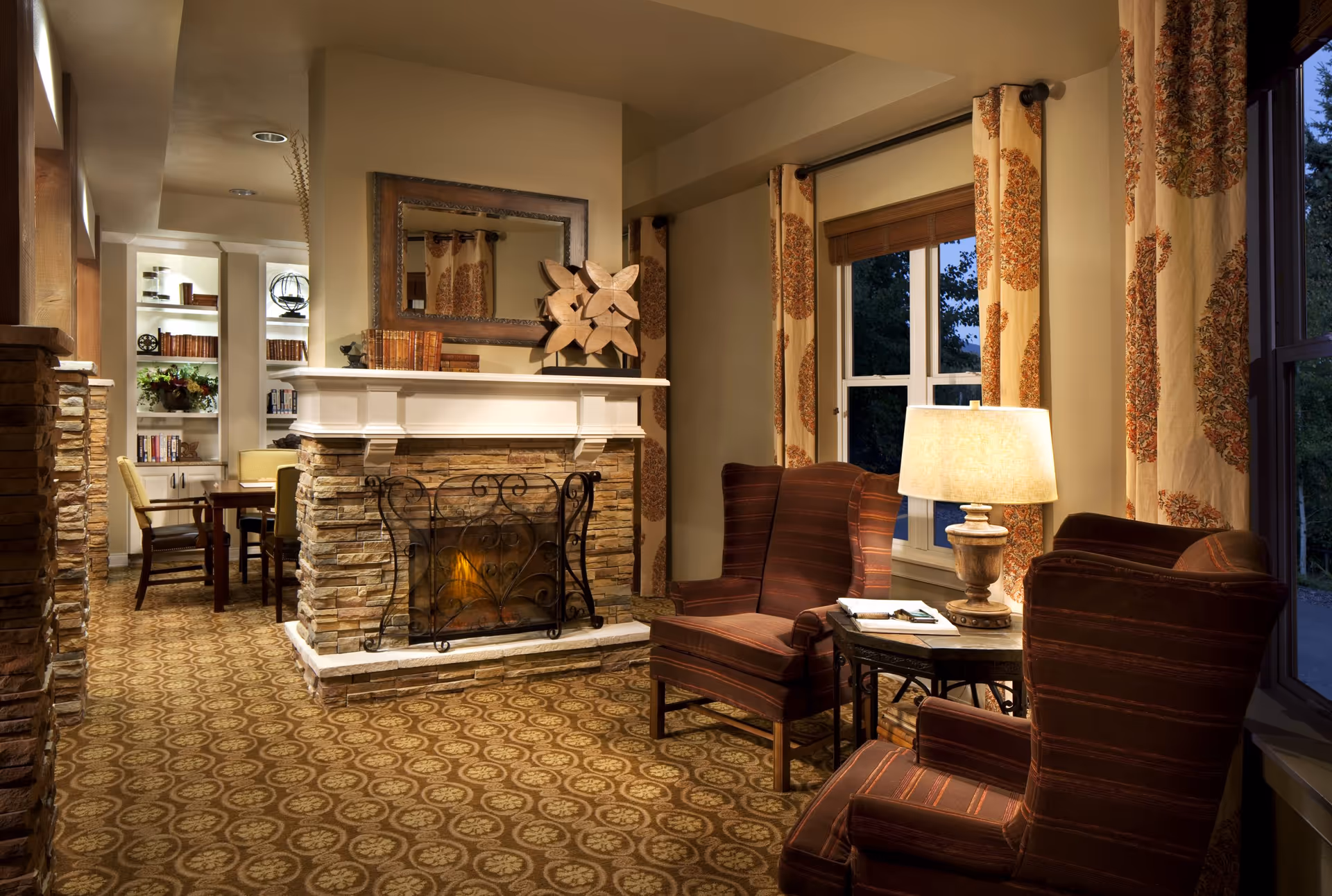 Cozy living room area with two upholstered armchairs and a side table with a lamp next to a window with patterned curtains. A stone fireplace with a decorative metal screen is centered in the room, topped with a large mirror, books, and a wooden sculpture. In the background, there is a small dining area with chairs and built-in shelves filled with books and decorative items.