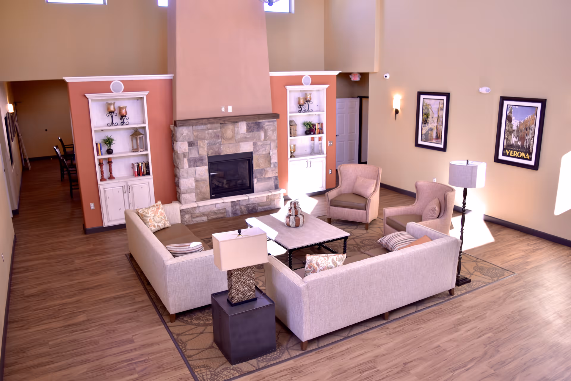 A cozy living room area in Brio Assisted Living featuring two beige sofas, two armchairs, a coffee table, a stone fireplace, two white shelves with decorative items, a floor lamp, and framed pictures on the wall. The room has wooden flooring and warm lighting.