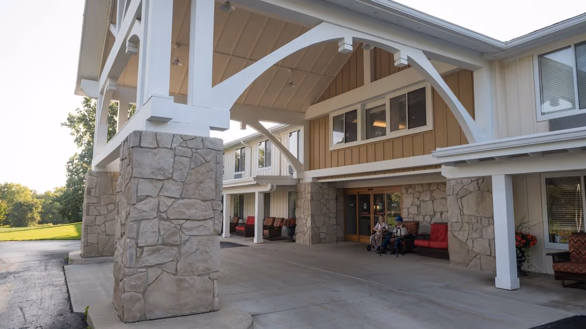 Covered entrance of a senior living facility with stone pillars, outdoor seating, and two people near the doorway.
