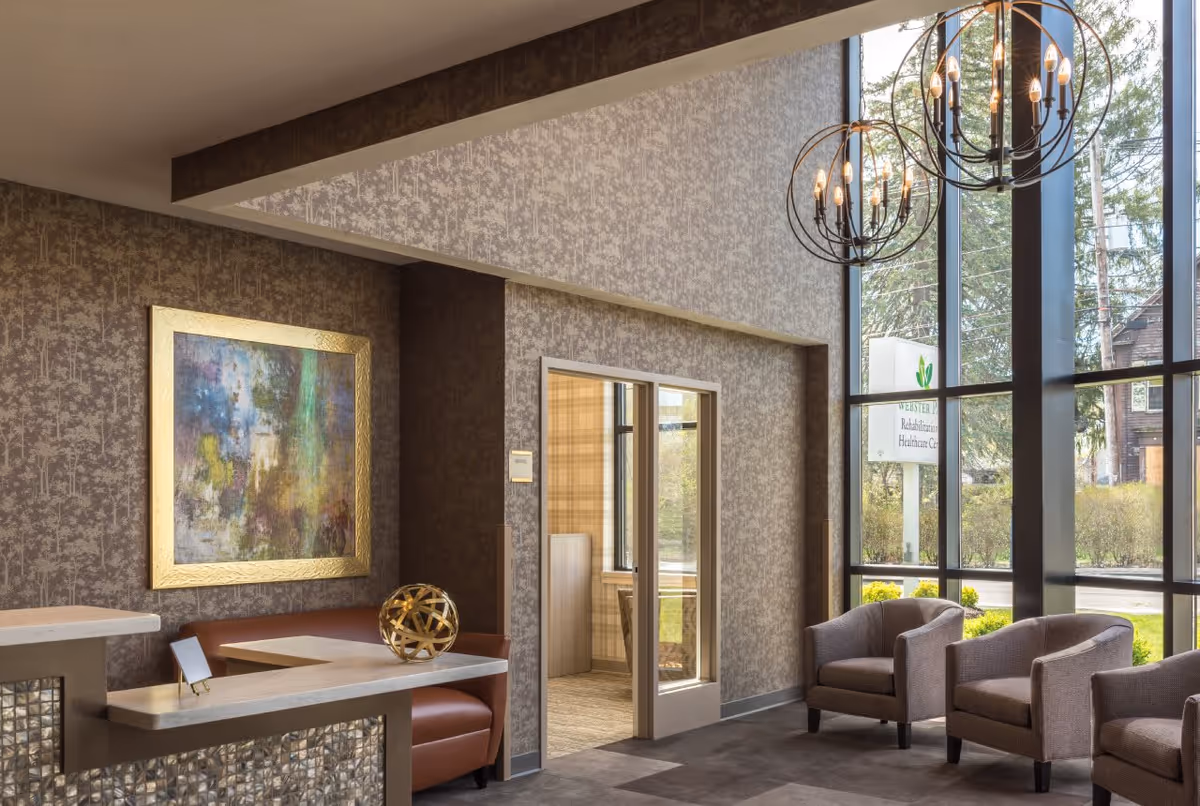 A modern and cozy interior seating area with three gray armchairs near large floor-to-ceiling windows. The windows reveal a sign outside that reads Webster Park Rehabilitation & Healthcare Center. The room features patterned wallpaper, a large abstract painting with a gold frame, a built-in bench with brown leather upholstery, and a decorative gold spherical sculpture on a white countertop. Two circular chandeliers with exposed bulbs hang from the ceiling.