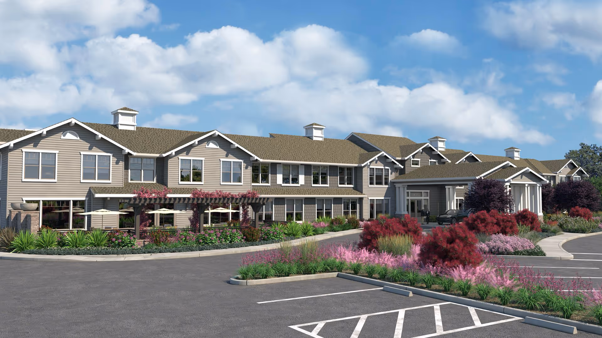 Exterior view of Oakmont of Silver Creek, a two-story senior living facility with beige siding and multiple windows. The building is surrounded by landscaped gardens with green shrubs and pink flowering plants. A paved driveway and parking area are visible in the foreground under a partly cloudy blue sky.