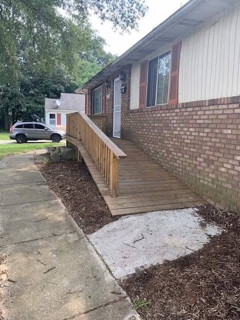 Exterior view of a single-story building with brick and siding walls, featuring a wooden ramp leading to a white door with a window. There are two windows with red shutters on the building. A gray car is parked on the driveway, and trees are visible in the background.