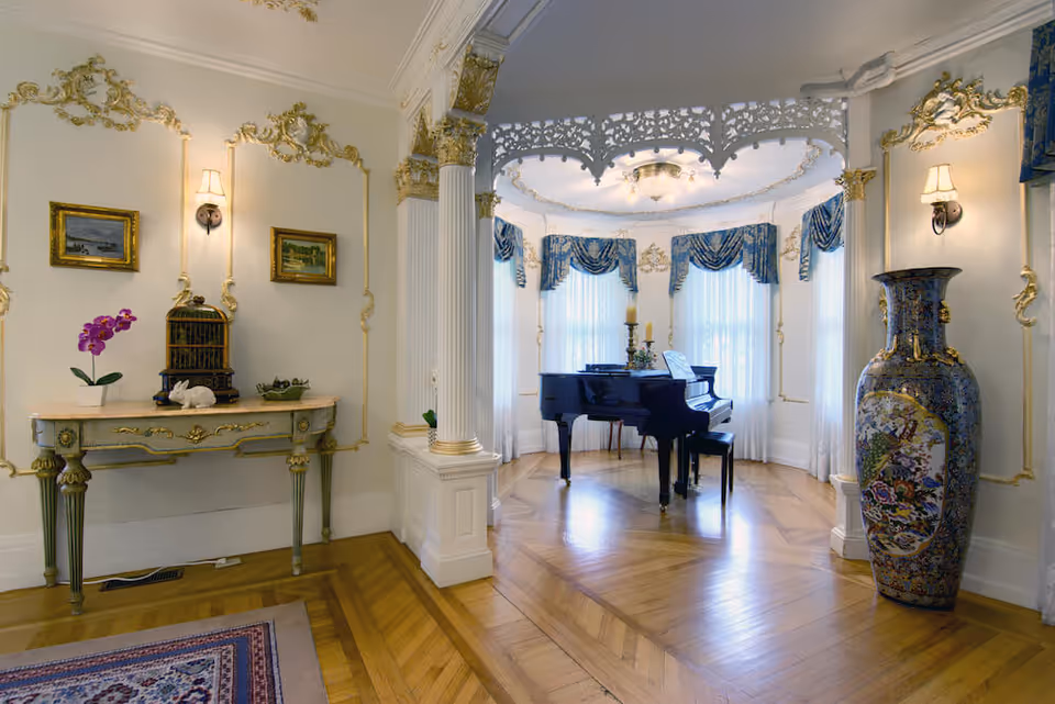 Elegant interior room with ornate white and gold trim, featuring a black grand piano in a bay window area with blue and gold drapes, a decorative large vase on the right, and a vintage console table with a purple orchid and small decorative items on the left.
