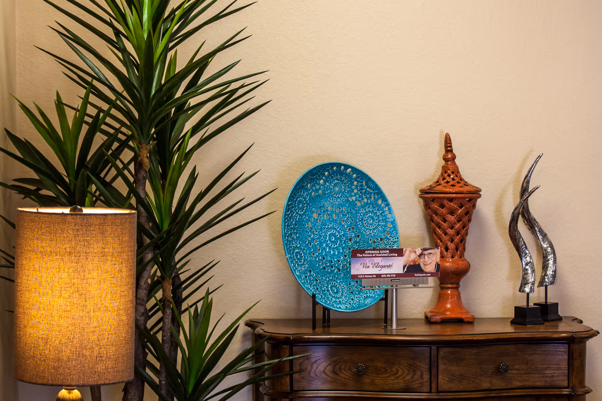 A wooden sideboard against a beige wall topped with a decorative turquoise plate, ceramic ornaments, a lamp, and a tall potted plant.