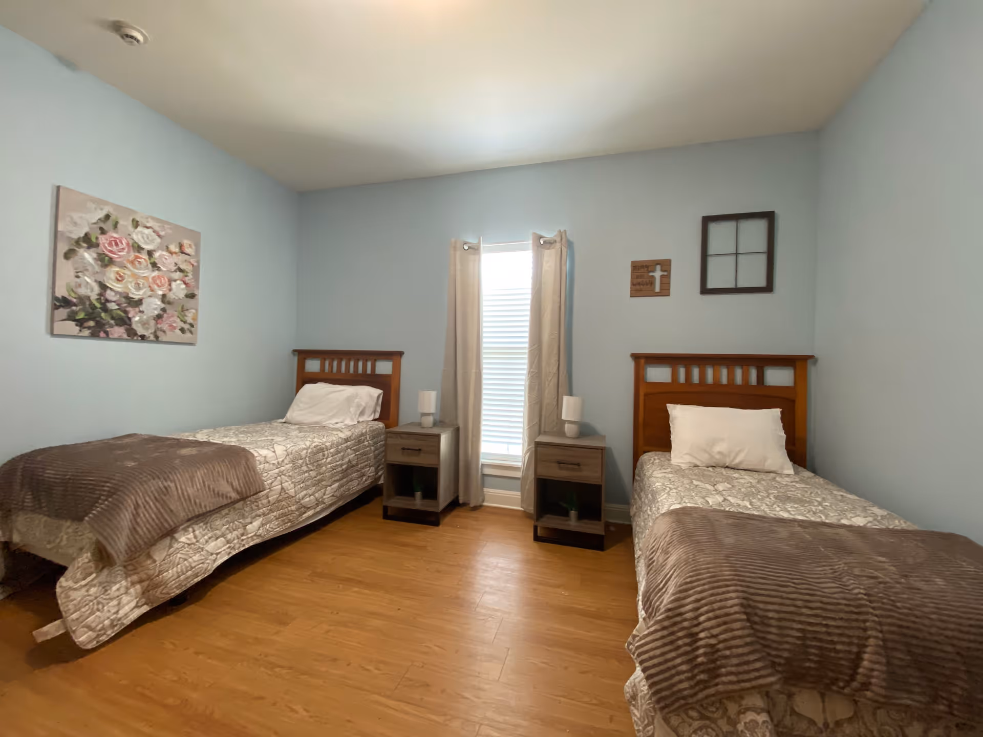A bedroom with two single beds separated by a window with beige curtains. Each bed has a wooden headboard, white pillow, and beige patterned bedding with a brown throw blanket. Between the beds are two matching nightstands, each with a small lamp and a decorative plant on the lower shelf. The walls are painted light blue and decorated with a floral painting on the left and two small wall hangings on the right.
