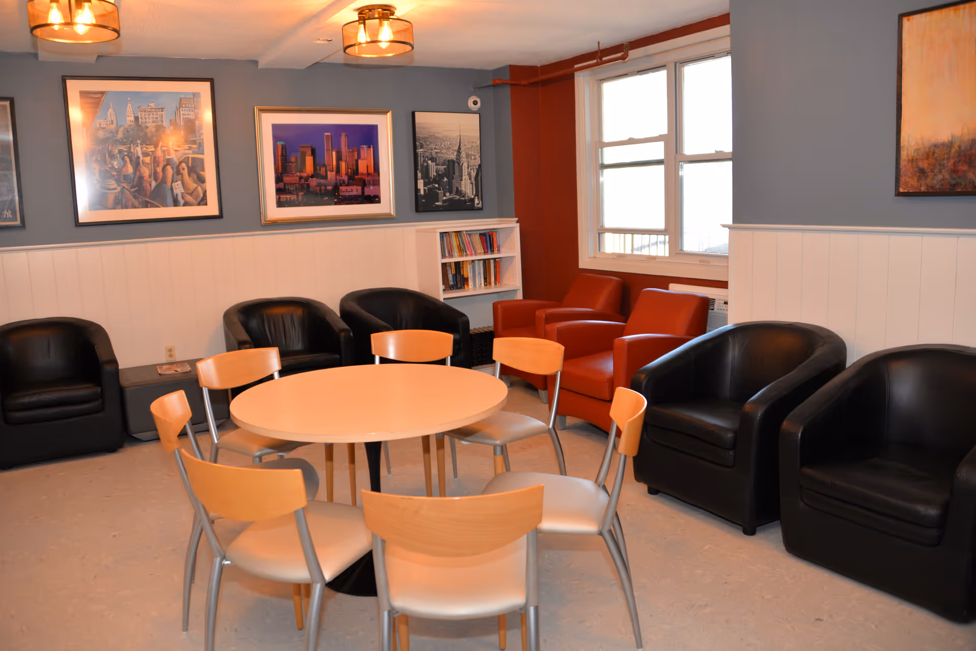 A cozy common area with a round wooden table surrounded by six chairs in the center. Around the room are several black and red armchairs. The walls are painted gray and white with framed cityscape artwork hanging. A window lets in natural light, and a small bookshelf with books is visible in the corner.