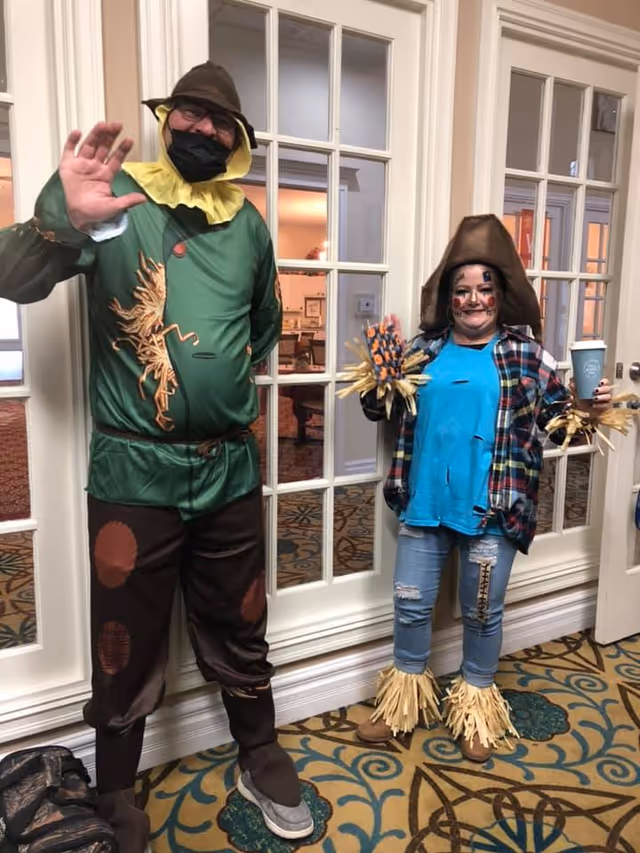 Two people dressed in scarecrow costumes standing indoors in front of glass-paneled doors. The person on the left is wearing a green and brown costume with a yellow collar and a black face mask, waving at the camera. The person on the right is wearing a blue shirt, plaid jacket, ripped jeans with straw details, and a brown hat, holding a cup and smiling with face paint resembling a scarecrow.