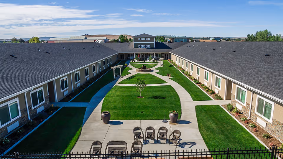 Aerial view of a senior living facility courtyard with a large green lawn, concrete walkways, outdoor seating with chairs and tables, potted plants, and a central fire pit area. The building surrounds the courtyard on three sides with multiple windows and a dark roof under a partly cloudy sky.