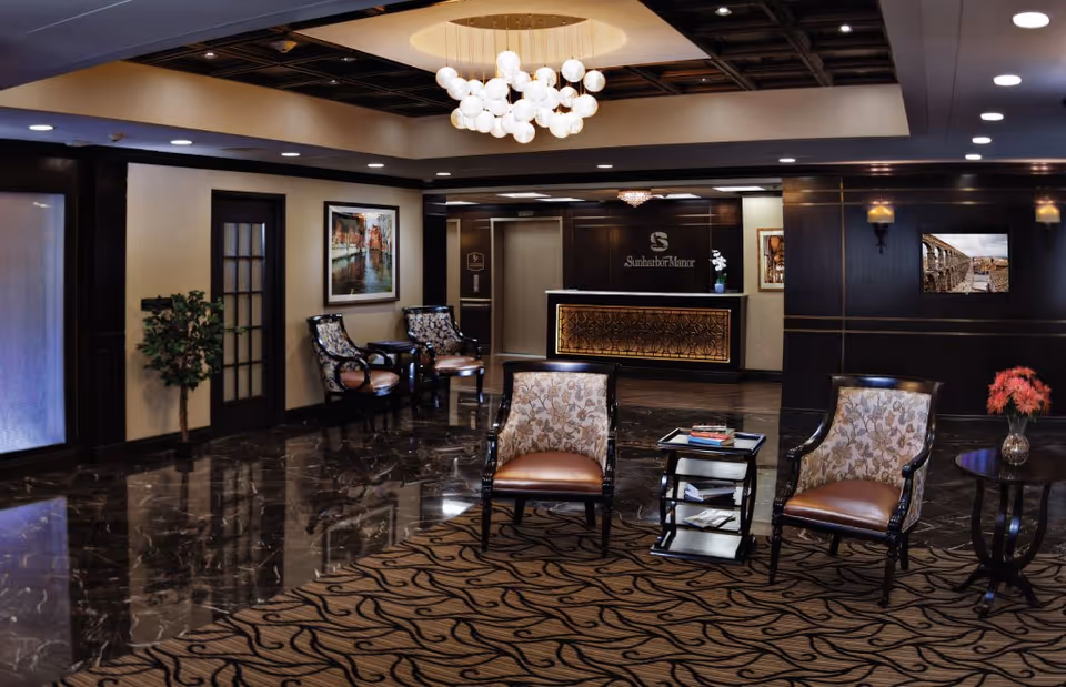 A spacious and elegant lobby area with polished dark marble floors and a patterned carpet. There are several upholstered chairs with floral patterns and leather seats arranged around small tables. The reception desk is visible in the background with the sign 'Sunharbor Manor' on the wall behind it. The ceiling features a modern chandelier with multiple round lights. The walls are decorated with framed artwork and sconces, and there is a potted plant near a door with frosted glass panels.