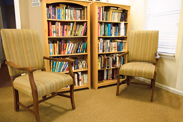 Two upholstered armchairs face each other in a carpeted reading nook in front of two wooden bookshelves filled with books.