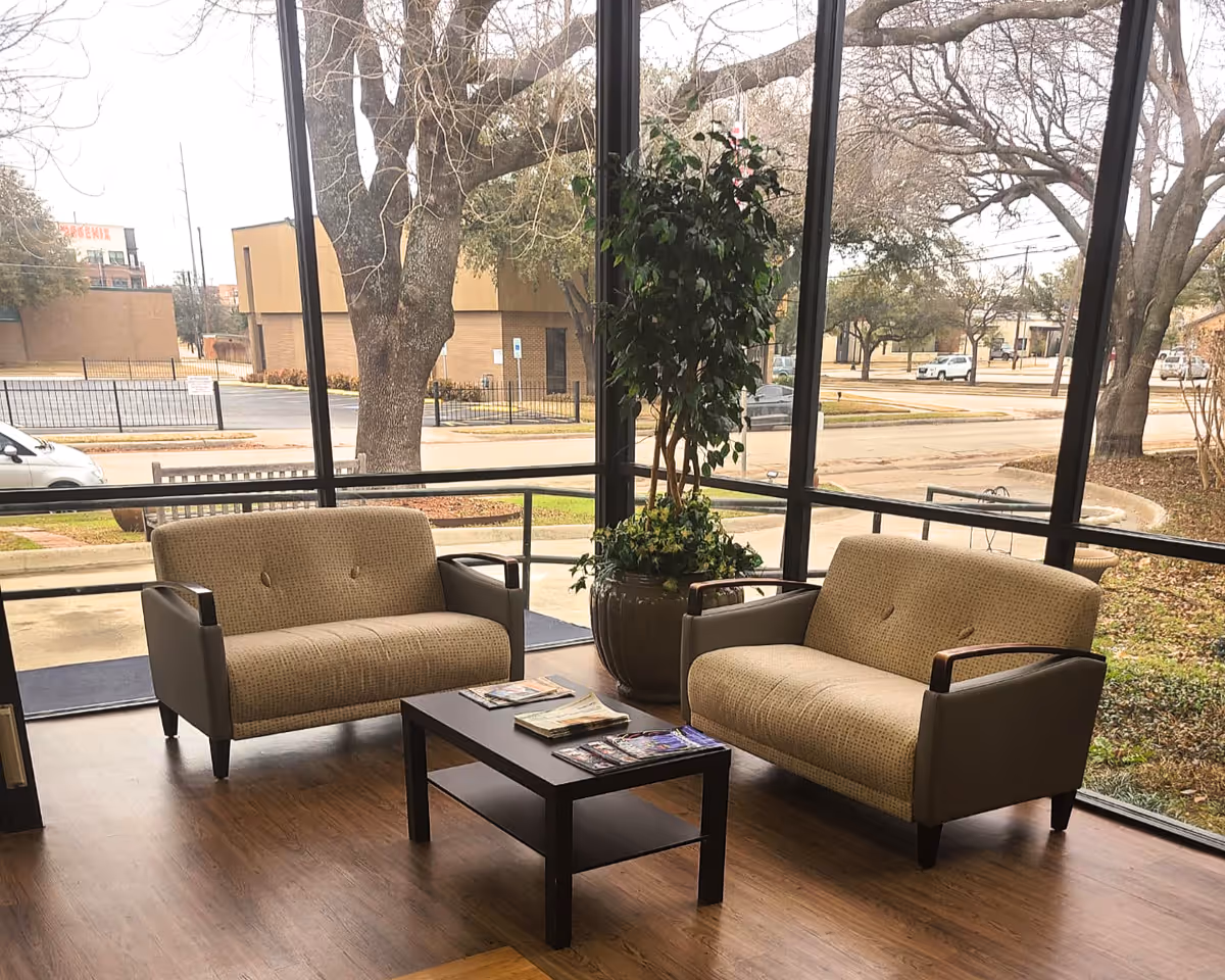 A seating area inside a building with two beige upholstered loveseats facing each other and a dark wooden coffee table between them. The area has large floor-to-ceiling windows showing an outside view of trees, a street, and parked cars. A large potted plant is placed behind the coffee table near the windows.