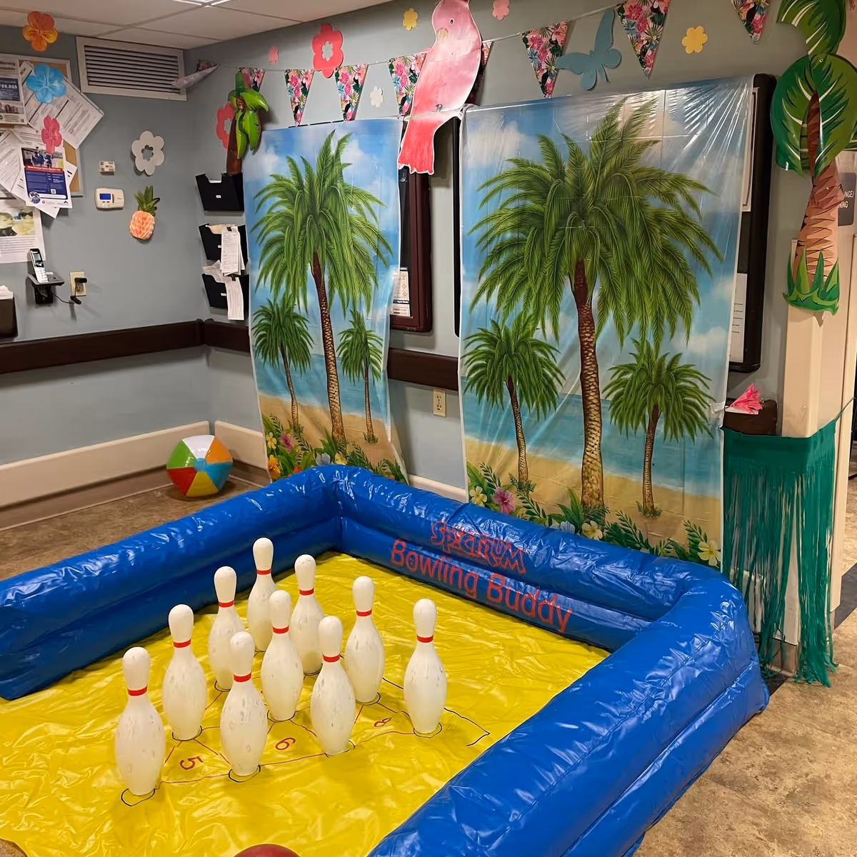 An indoor recreational area with an inflatable bowling game set up on the floor. The bowling pins are arranged on a yellow mat with a blue inflatable border labeled 'Spectrum Bowling Buddy.' The walls are decorated with tropical-themed decorations including palm tree backdrops, paper flowers, a beach ball, and a pink bird cutout hanging from a string of triangular flags.