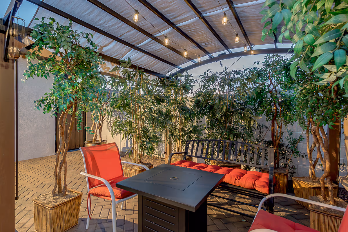 Outdoor covered patio area with a black metal bench with red cushions, two red cushioned chairs, a black square table, potted plants, and string lights hanging from the ceiling.