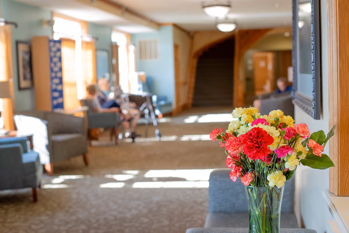 A bright and spacious assisted living facility common area with a vase of colorful flowers in the foreground. In the background, there are several armchairs and elderly residents sitting and conversing near large windows that let in natural light. The area has carpeted flooring and wooden accents, with a staircase visible further back.