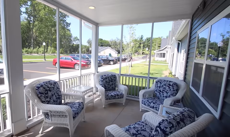 Screened front porch with white wicker chairs and blue patterned cushions overlooking a parking lot and lawn.