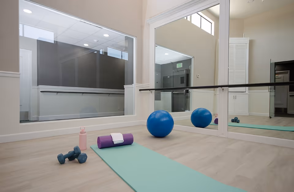Bright exercise/yoga room with a mat, dumbbells, foam roller, water bottle and an exercise ball in front of a mirrored wall.