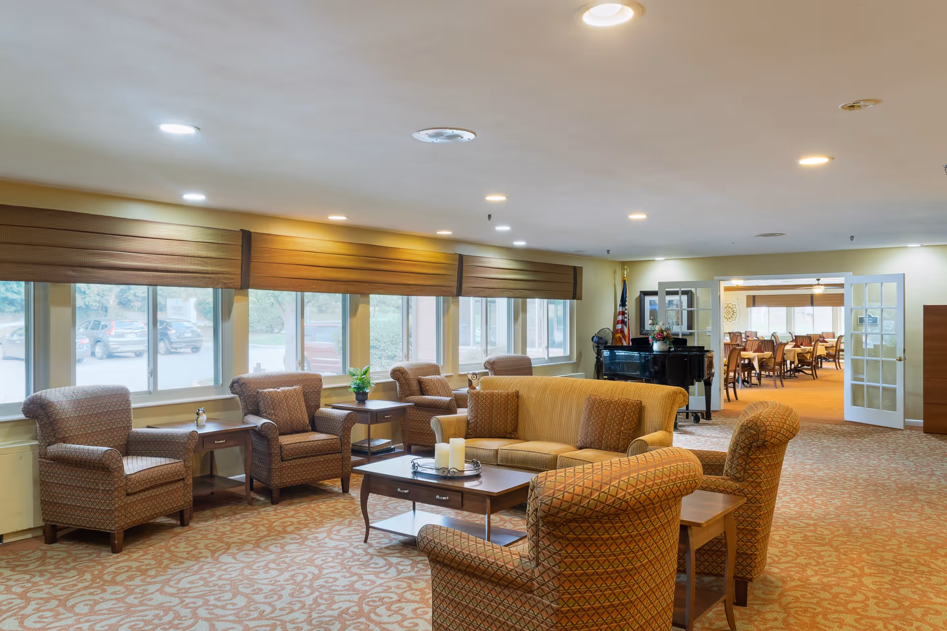 A spacious and well-lit living room area in Deer Park Assisted Living featuring multiple patterned armchairs and a sofa arranged around a wooden coffee table with candles. Large windows with brown blinds let in natural light. In the background, there is a black piano with a flower arrangement on top and an American flag beside it. Double glass doors lead to a dining room with tables and chairs.